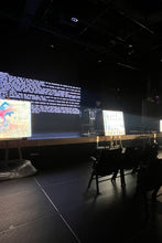 Load image into Gallery viewer, Dark room with stage setup and screens displaying text and graphics